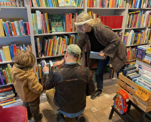 Interiör från bokhandeln där kvinna visar en bok medan liten flicka och hennes pappa tittar på framför belamrad bokhylla
