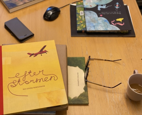 Bild på boken Efter stormen, en bilderbok med starkt gul färg och ett rött flygplan, som ligger på ett skrivbord där det också ligger glasögon och står en kaffemugg