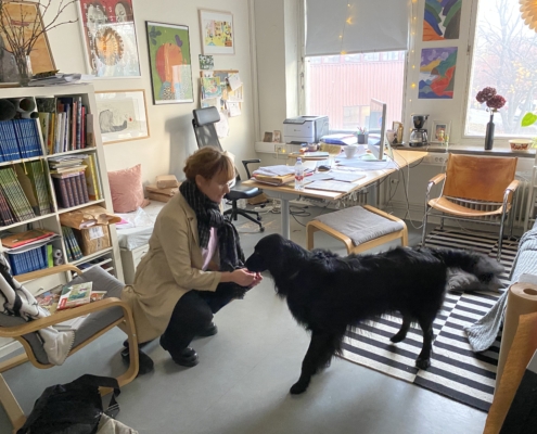 Bild på ett rum med bokhylla och skrivbord och massa papper och i mitten på golvet sitter en tjej på huk och klappar en svart hund
