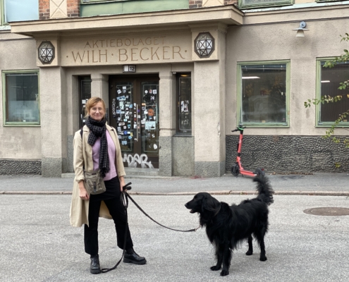 Bild på en tjej i byxor och beige rock som står med sin svarta hund utanför en port där det står Aktiebolaget Wilh. Becker ovanför dörren