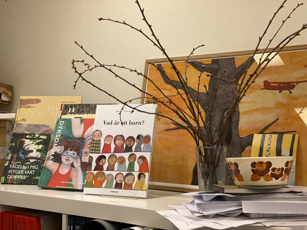Bokhylla som visar olika färglada bilderboksböcker och en hundskål och en illustration med träd och flygplan i bakgrunden