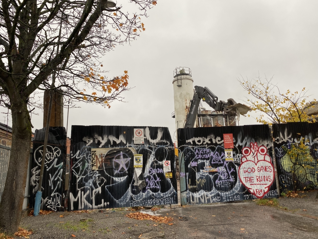 Ett stort svart plank med färggrann graffiti och bakom det härjar en grävskopa runt