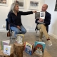 Bild på en man och en kvinna som sitter på varsin stol vid en vägg med tavlor och samtalar om en bok som mannen håller i handen