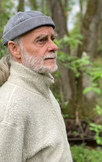 Bild på Stefan Casta i blå mössa stående framför suddig rak trädstam och seende lite sorgsen ut iklädd gråvit fleecejacka