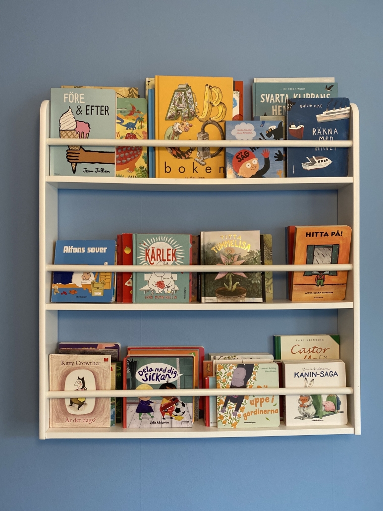Vit barnbokhylla på blå vägg, i hyllan syns en massa olika småbarnsböcker