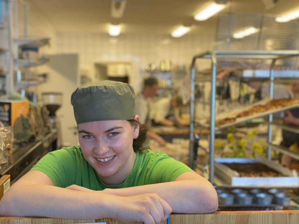 Bild på tjej i grön t-shirt som ler med grön kockmössa på huvudet och i bakgrunden bakplåtar och ett kondiskök