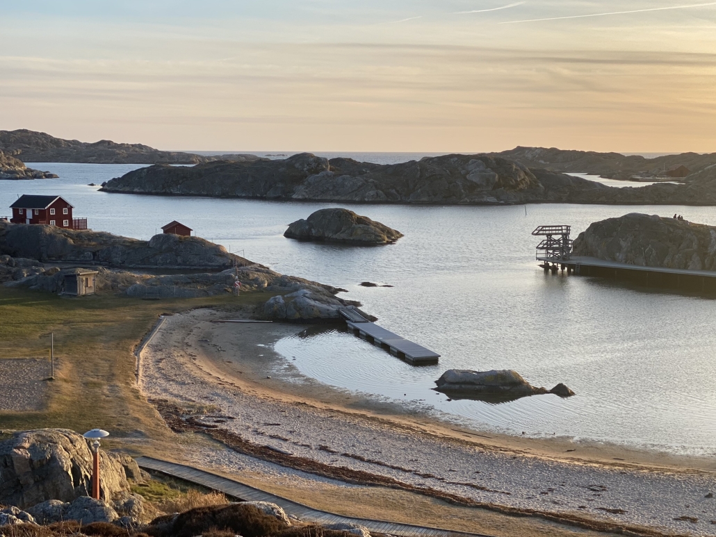 Bild på lagunliknande strand med klippöar utanför och en röd byggnad i en hörnet av stranden, ett gyllene skymningsljus vilar över havet och stranden