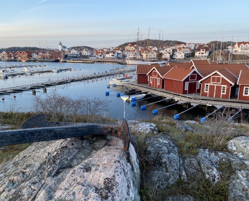 Bild på fiskesamh'ället Sk'ärhamn visar ett ankare på klippa i förgrunden och i bakgrunden syns röda sjöbodar vid vatten och längre bort hela samhället med sina vita skepparhus och en vit kyrka nedanför grå berg