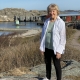 Bild av Benita Nilsson i helfigur med vit skjorta och svarta byxor och i bakgrunden den röda avlånga museibyggnaden som är Nordiska akvarellmuseetvid klippor och vatten