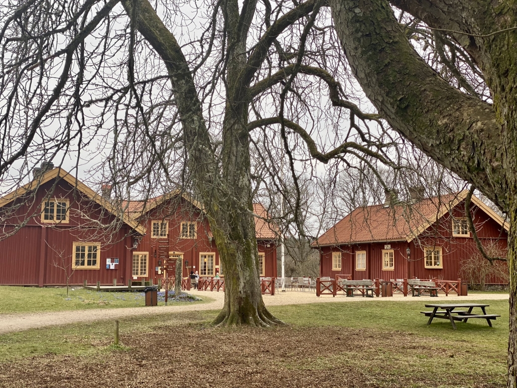 Bild på tre röda herrgårdsliknande byggnader med gulfärgade fönsterramar och med kaffebord i förgrunden inramat av svarta träd och trädstammar